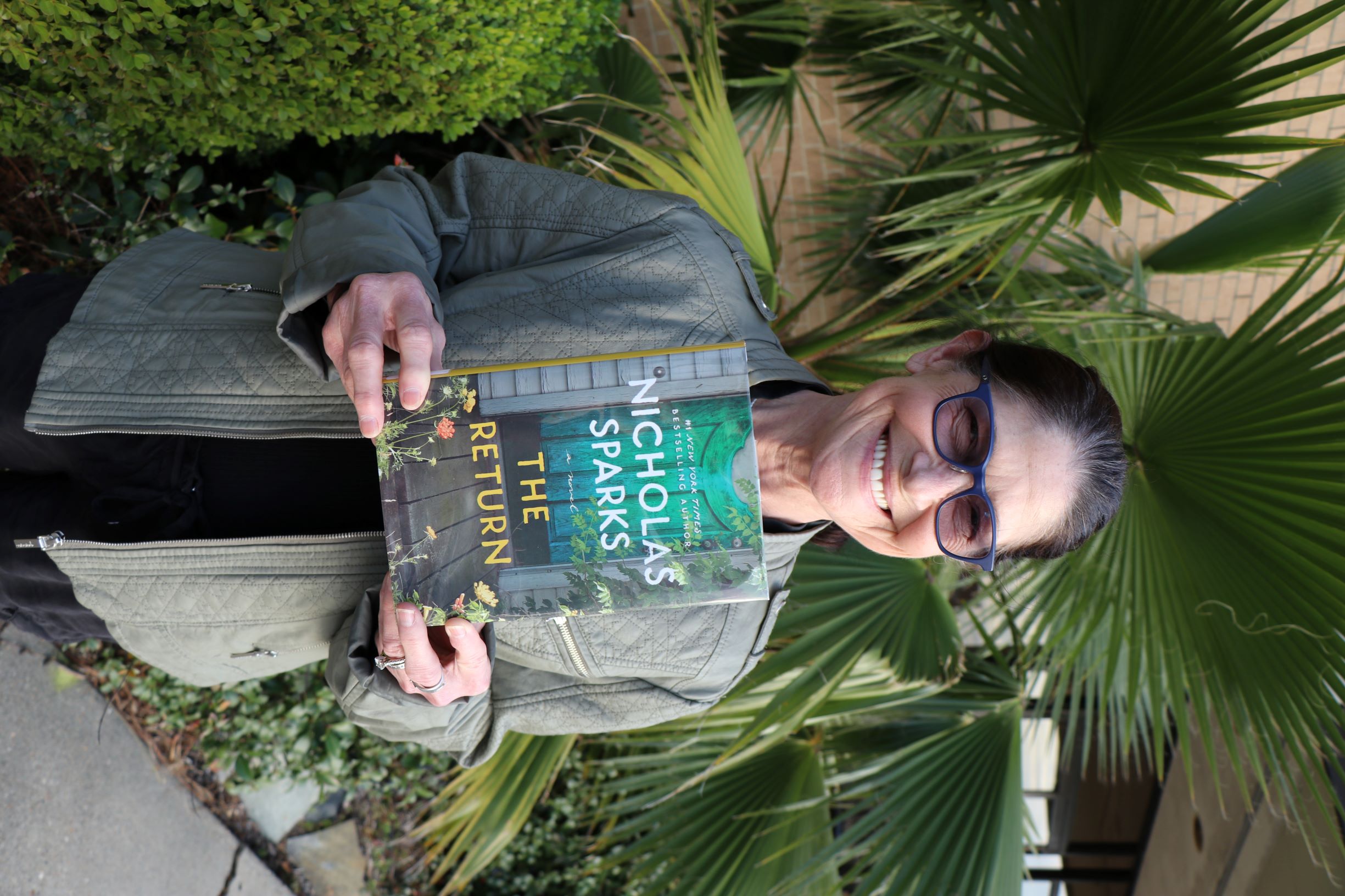 A woman stands in front of a plant. She is holding the book The Return.