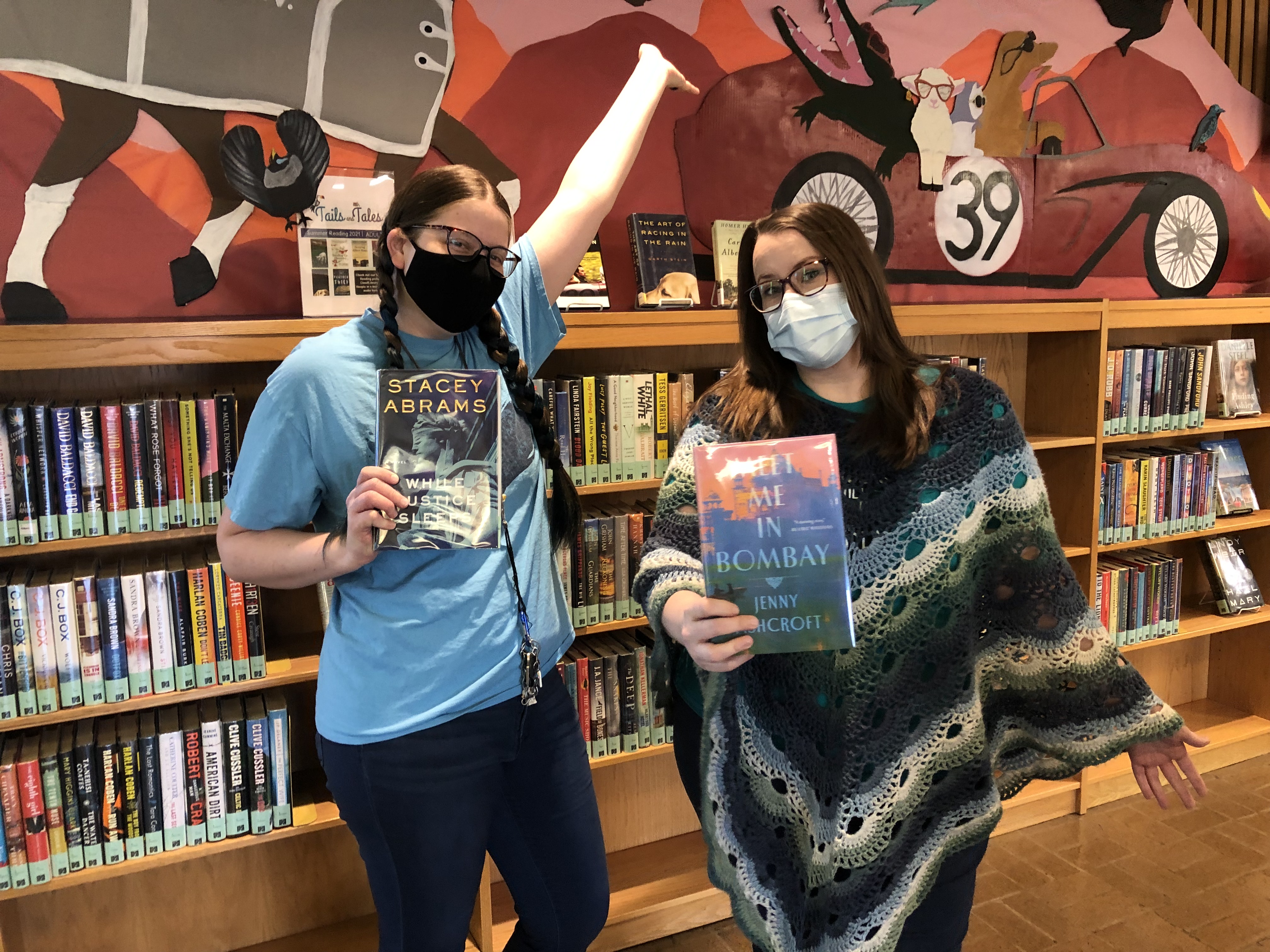 Two women wearing masks and holding books stand in front of a book shelf.