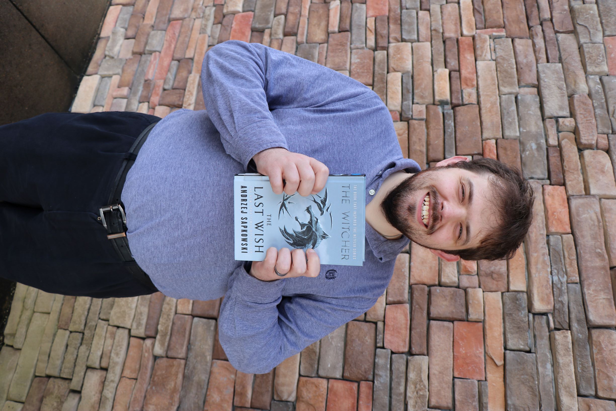 A man stands in front of a brick wall. He is holding the book The Last Wish.