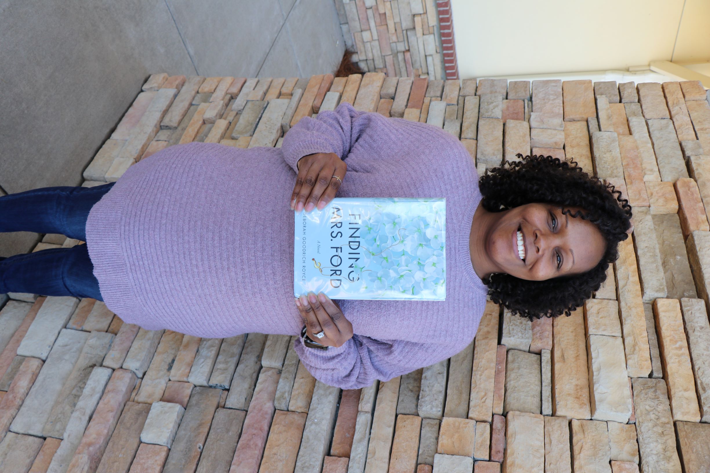 A woman stands in front of a brick wall. She is holding the book Finding Mrs. Ford